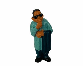 May include: A small figurine of a man wearing a light blue shirt, dark blue trousers, and black shoes. He is wearing sunglasses and has his hand on his chin, as if he is thinking.
