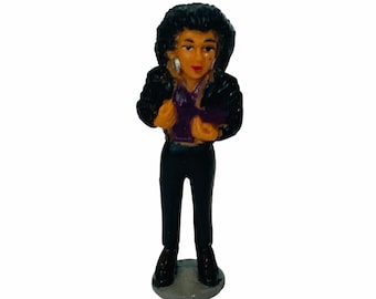 May include: A small plastic figurine of a person wearing a black leather jacket, purple shirt, and black trousers. The figurine has black hair and brown eyes.