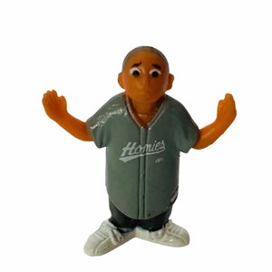 May include: A gray plastic figurine of a person wearing a gray baseball jersey with the word "Homies" printed on it. The figurine has brown skin and is wearing white sneakers.