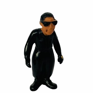 May include: A black plastic figurine of a person wearing a black jumpsuit, sunglasses, and gloves.