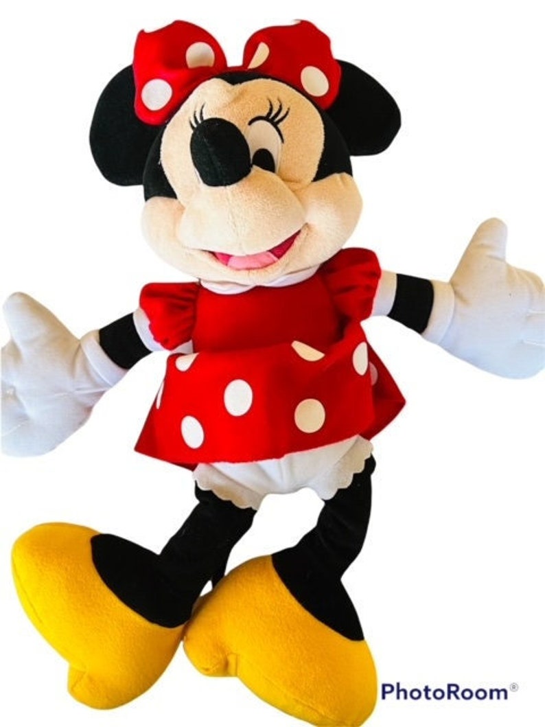 May include: A plush toy of Minnie Mouse wearing a red dress with white polka dots and a red bow with white polka dots. She has black eyes, a black nose, and a big smile. She is standing on her two yellow feet.