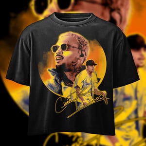 May include: Black t-shirt with a graphic of a man wearing sunglasses, set against a yellow and orange backdrop. The shirt features the signature "Chris Brown" in yellow script.
