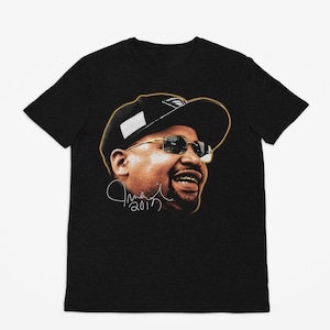 Puede incluir: Camiseta negra con un retrato de un hombre con gafas de sol y una gorra de béisbol. El retrato tiene un contorno dorado e incluye una firma con el año 2017. El hombre tiene dientes de oro.