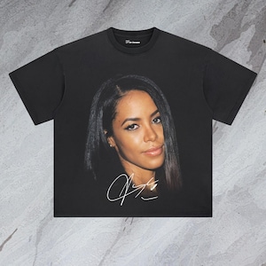 May include: Black t-shirt featuring a printed image of a woman with long dark hair and a white signature. The signature reads "Aaliyah".