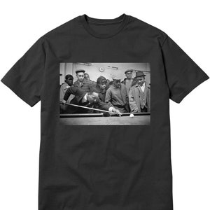 May include: Black and white photograph of a man playing pool with a group of young men watching. The man is wearing a suit and tie, and he is holding a pool cue. The young men are wearing casual clothing.