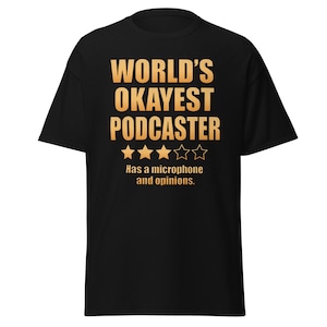 May include: A black short-sleeved t-shirt featuring the gold text "WORLD'S OKAYEST PODCASTER" above three filled-in stars and two empty stars. Below the stars, the text reads "Has a microphone and opinions." in a matching gold colour.