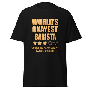 May include: Black t-shirt with the text "WORLD'S OKAYEST BARISTA" in gold lettering. Below the text are four gold stars and the phrase "Spelled my name wrong. Twice... It's Bob."
