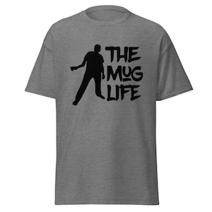 Puede incluir: Una camiseta gris jaspeado con un gráfico negro. El gráfico presenta la silueta de una persona sosteniendo una taza, con las palabras "THE MUG LIFE" en una fuente audaz y estilizada. La camiseta tiene mangas cortas y cuello redondo.