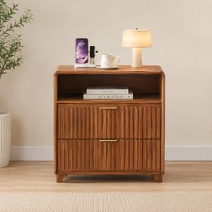 May include: A wooden nightstand with two drawers and a shelf. The nightstand has a warm brown finish and vertical ribbed detailing on the drawer fronts. A lamp, books, a mug, and a phone are on top. The nightstand is 28 inches tall.
