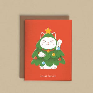 Puede incluir: Tarjeta de Navidad con un gato blanco disfrazado de árbol de Navidad, adornado con adornos y una estrella. La tarjeta tiene un fondo rojo con el texto "FELINE FESTIVE" y está sobre un fondo neutro.