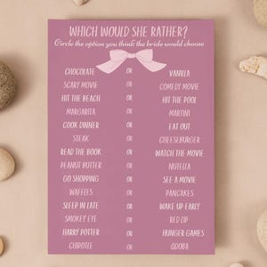 Puede incluir: Una tarjeta de juego de despedida de soltera de color malva con el texto "WHICH WOULD SHE RATHER?" y una lista de opciones. La tarjeta está rodeada de piedras lisas y de color claro sobre una superficie beige.