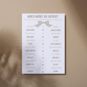 Puede incluir: Una tarjeta blanca con el texto "WHICH WOULD SHE RATHER?" y una lista de opciones, incluyendo "Chocolate o Vainilla" y "Película de miedo o Comedia". Un lazo marrón claro está en la parte superior.