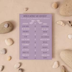 Puede incluir: Una tarjeta de juego de despedida de soltera lavanda con el texto "Which Would She Rather?" está rodeada de piedras lisas de color claro sobre una superficie beige. La tarjeta presenta un gráfico de lazo y una lista de opciones.