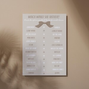 Puede incluir: Una tarjeta gris claro con el texto "WHICH WOULD SHE RATHER?" y una lista de opciones. Las opciones incluyen chocolate o vainilla, película de miedo o comedia, y más. Un lazo decorativo está en la parte superior.