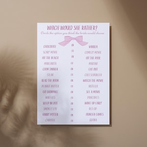 Puede incluir: Una tarjeta blanca con texto rosa y un lazo rosa. El texto dice "WHICH WOULD SHE RATHER?" seguido de una lista de opciones para un juego de despedida de soltera. La tarjeta está sobre un fondo neutro.