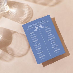 Puede incluir: Una tarjeta de juego azul "Which Would She Rather?" con un lazo, con varias opciones como "Chocolate" y "Ver una película". La tarjeta está sobre una superficie marrón, con dos vasos de agua en el fondo.