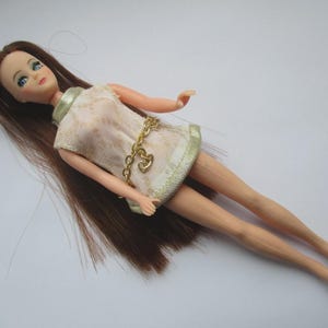 May include: A vintage doll with long brown hair and blue eyes. The doll is dressed in a cream-coloured dress with gold trim and a gold chain belt with a heart-shaped charm. The doll is lying down on a white surface.