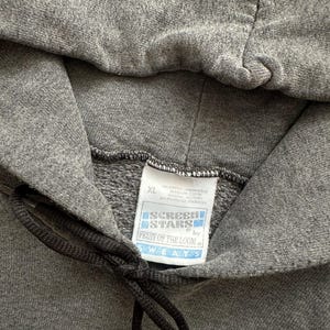 Puede incluir: Primer plano de una sudadera con capucha gris con cord&oacute;n. La etiqueta dice "SCREEN STARS by FRUIT OF THE LOOM SWEATS" e indica una talla XL. La tela parece ser un material suave y de peso medio.