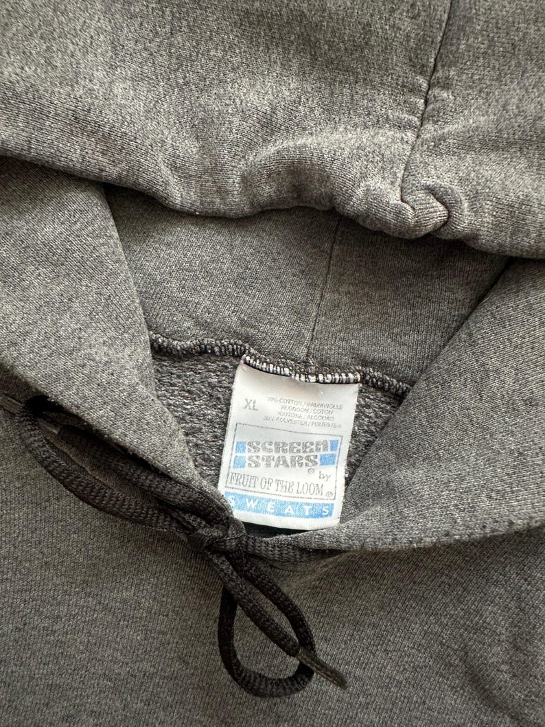 Puede incluir: Primer plano de una sudadera con capucha gris con cord&oacute;n. La etiqueta dice "SCREEN STARS by FRUIT OF THE LOOM SWEATS" e indica una talla XL. La tela parece ser un material suave y de peso medio.
