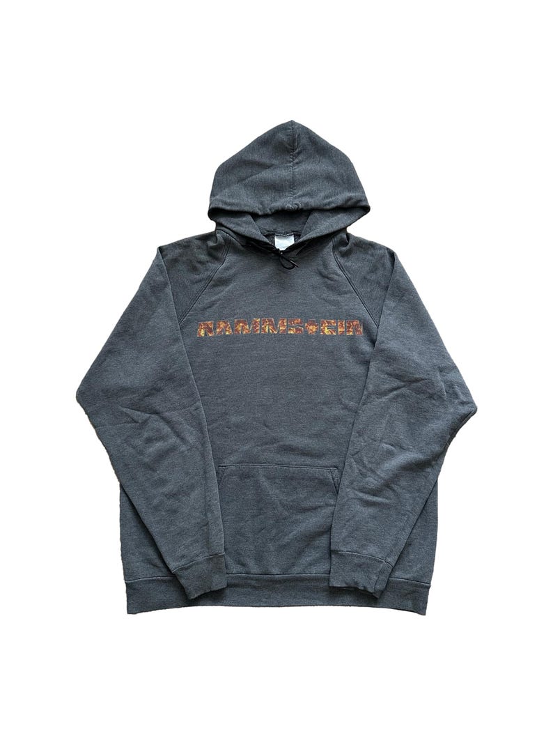 Puede incluir: Una sudadera con capucha gris oscuro con la palabra "RAMMSTEIN" impresa en el pecho en un dise&ntilde;o texturizado y multicolor. La sudadera tiene un bolsillo delantero y una capucha con cord&oacute;n. La prenda est&aacute; hecha de un material suave.