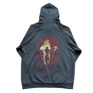 Puede incluir: Sudadera con capucha gris oscuro con un gr&aacute;fico en la espalda. El gr&aacute;fico presenta una figura en blanco y dorado sobre un fondo rojo. La sudadera con capucha est&aacute; hecha de un material suave.