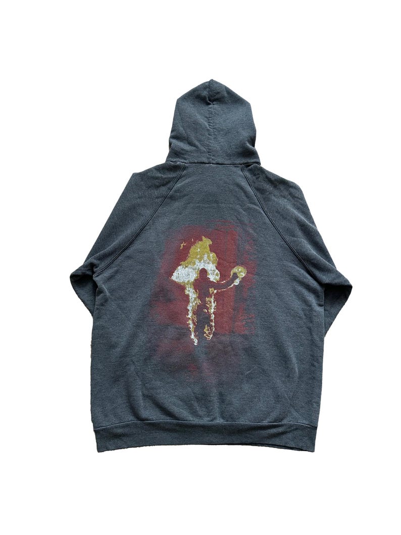 Puede incluir: Sudadera con capucha gris oscuro con un gr&aacute;fico en la espalda. El gr&aacute;fico presenta una figura en blanco y dorado sobre un fondo rojo. La sudadera con capucha est&aacute; hecha de un material suave.