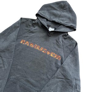 Puede incluir: Sudadera con capucha gris oscuro con bolsillo delantero y capucha con cord&oacute;n. La palabra "RAMMSTEIN" est&aacute; impresa en el pecho con un dise&ntilde;o llameante naranja y amarillo. La sudadera con capucha est&aacute; hecha de un material suave y c&oacute;modo.