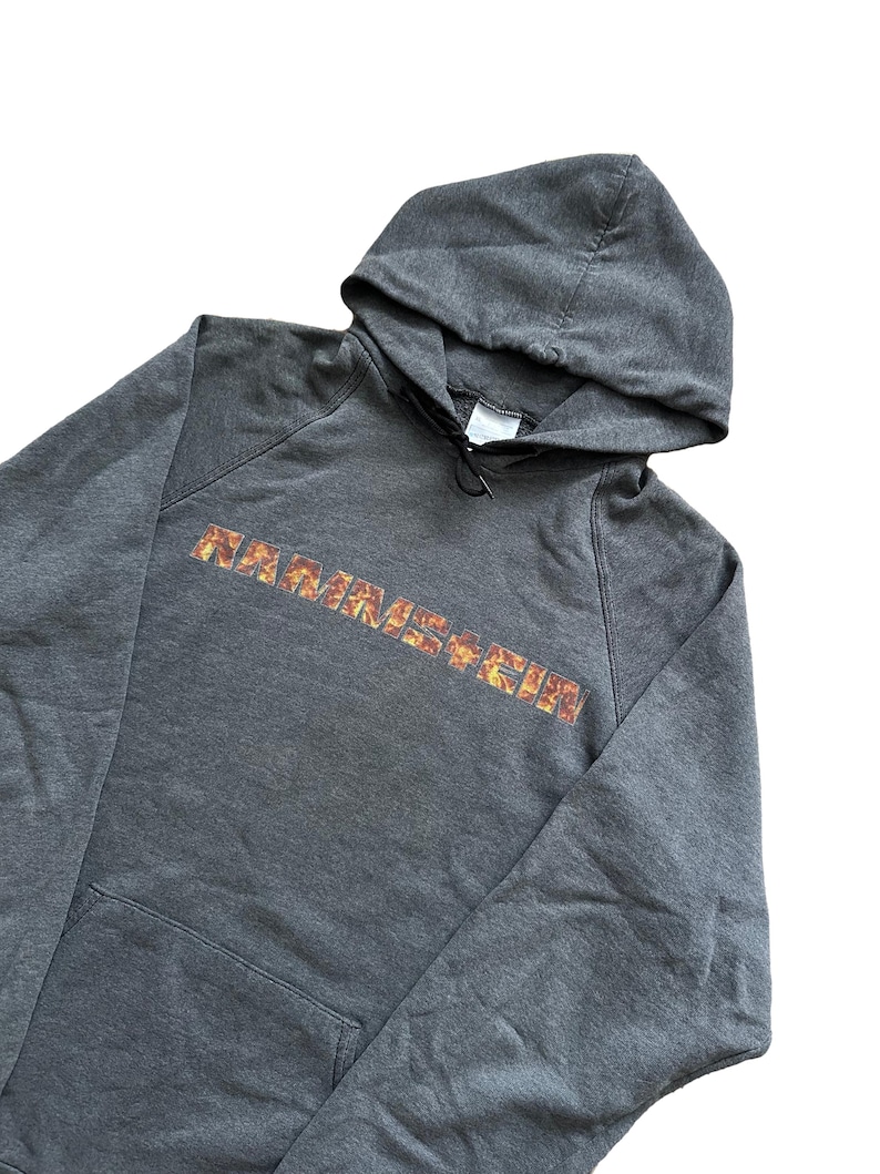 Puede incluir: Sudadera con capucha gris oscuro con bolsillo delantero y capucha con cord&oacute;n. La palabra "RAMMSTEIN" est&aacute; impresa en el pecho con un dise&ntilde;o llameante naranja y amarillo. La sudadera con capucha est&aacute; hecha de un material suave y c&oacute;modo.
