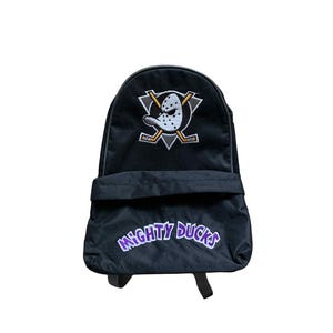 Op de afbeelding: Zwarte rugzak met de tekst Mighty Ducks in paars en een logo van een hockeymasker en gekruiste hockeysticks. Hij heeft een voorvak en verstelbare bandjes, ideaal voor het dragen van boeken en uitrusting.