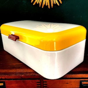 Peut inclure: Une boîte à pain en émail blanche et jaune avec une poignée marron. Le couvercle a un motif floral blanc.