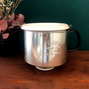 May include: A silver metal coffee filter with a white plastic lid and a black handle. The filter has the text "Melitta Schnell-Filter No 103 Für 3-15 Tassen" printed on the side.