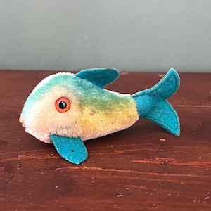May include: A soft toy fish with a teal and yellow body, teal fins, and a round orange eye. The fish is lying on a dark brown wooden surface.