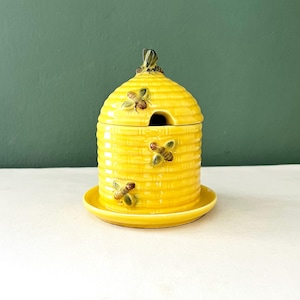 Può includere: Un vasetto di miele in ceramica gialla a forma di alveare, completo di coperchio e piattino. Il vaso è decorato con api tridimensionali. Il coperchio ha una piccola apertura per un cucchiaio. L'alveare è appoggiato su un piattino giallo.