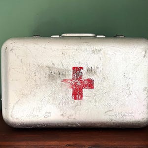 Op de afbeelding: Een vintage EHBO-doos van zilverkleurig metaal met een rood kruis op de voorkant.