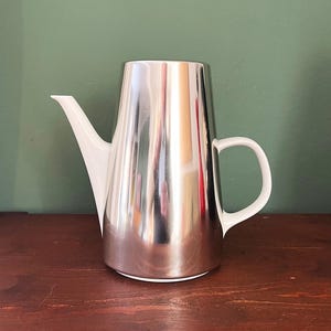 Puede incluir: Una cafetera de acero inoxidable con boquilla y asa blancas. La cafetera tiene una superficie pulida y reflectante y una forma cónica. El asa tiene un diseño simple y curvado.
