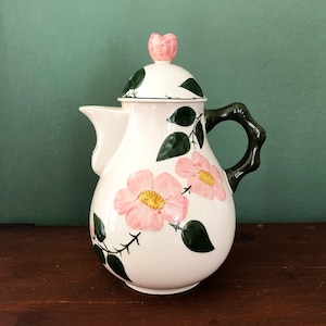 Può includere: Teiera in ceramica bianca con un design floreale con rose rosa e foglie verdi. La teiera ha un coperchio con una piccola rosa rosa in cima e un manico marrone.