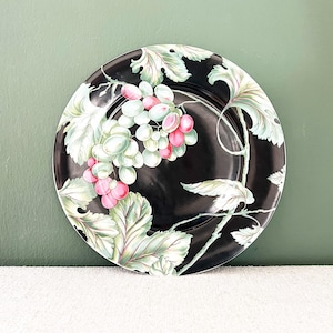 Peut inclure: Une assiette décorative ronde noire avec un motif de raisin. L'assiette présente une grappe de raisins roses et verts, entourée de feuilles et de vignes vertes et blanches. L'assiette est posée sur un fond vert.