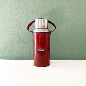 Può includere: Un thermos per caffè rosso con coperchio argentato e manico in pelle. Il thermos ha una fascia al centro con la scritta "COFFEE" stampata. Lo sfondo è verde tenue.