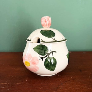 Può includere: Zuccheriera in ceramica color crema con coperchio e fessura per cucchiaio. Decorata con fiori rosa e foglie verdi. Il coperchio ha una maniglia a forma di rosa rosa.