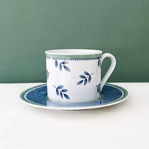 May include: A white ceramic teacup and saucer set. The cup and saucer have a blue and green leaf design. The cup has a handle and sits on a matching saucer. The set is against a green backdrop.
