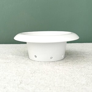 Peut inclure: Un pot de plante en céramique blanche avec un large bord plat et une base cylindrique. La base comporte plusieurs petits trous de drainage. Le pot est posé sur une surface claire et un fond vert.