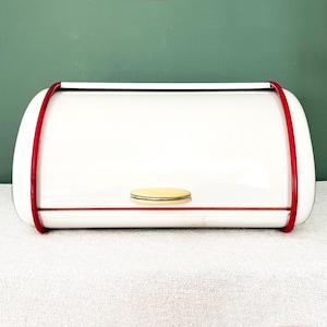 Può includere: Un portapane bianco con finiture rosse e un piccolo pomello rotondo color oro. Il portapane ha una parte superiore curva ed è posto su uno sfondo verde. È un articolo per la conservazione in cucina.