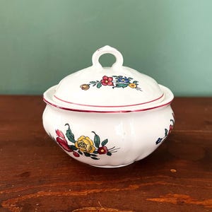 Pode incluir: Um açucareiro de cerâmica branca com tampa, apresentando um design floral em tons de vermelho, amarelo e azul. A tampa tem uma pequena alça.