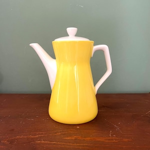 Puede incluir: Una cafetera vintage de cerámica amarilla y blanca. El cuerpo de la cafetera es de color amarillo brillante, con un pico, asa y tapa blancos. La cafetera tiene un diseño clásico y aerodinámico, ideal para servir café o té.