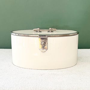 Op de afbeelding: Crèmekleurige, ovale container met een zilverkleurige handgreep en sierlijsten. De container heeft een scharnierend deksel en een sluiting. De achtergrond is effen groen.