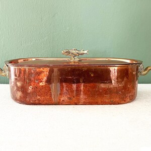 Può includere: Una pirofila rettangolare in rame con coperchio. La pirofila ha manici in ottone e una maniglia a forma di pesce sul coperchio. Il rame ha una patina rosso-marrone e le maniglie sono dorate. La pirofila è su una superficie bianca.