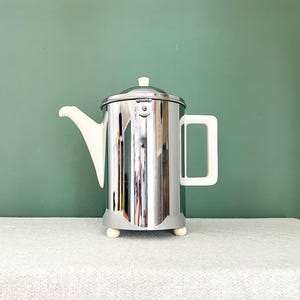 Könnte beinhalten: Eine Vintage-Kaffeekanne aus Chrom mit weißem Griff, Ausguss und Deckelknopf. Die Kanne hat einen zylindrischen Körper und steht auf einem grauen Sockel mit drei kleinen weißen Füßen. Der Hintergrund ist einfarbig türkis.