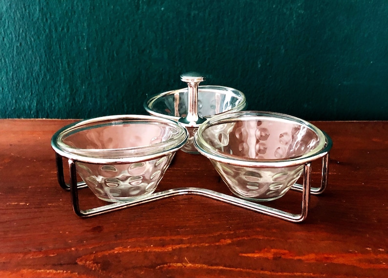 K&ouml;nnte beinhalten: Ein silberfarbenes Serviergeschirr mit drei durchsichtigen Glasschalen. Jede Schale hat ein strukturiertes Muster und wird von einem silbernen Metallrahmen gehalten. Der zentrale Griff ist ebenfalls silberfarben. Der Hintergrund ist dunkelgr&uuml;n.
