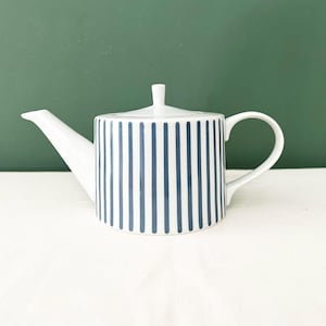 Può includere: Teiera in ceramica bianca con coperchio e manico. La teiera è decorata con strisce blu verticali. Il beccuccio è lungo e sottile. Lo sfondo è di colore verde unito.