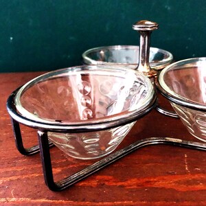 K&ouml;nnte beinhalten: Ein Vintage-Gew&uuml;rzset mit drei durchsichtigen Glasschalen mit strukturiertem Muster, die jeweils in einem silberfarbenen Metallrahmen gehalten werden. Ein zentraler Griff verbindet sich mit einer kleineren, runden Glasschale. Das Set steht auf einer Holzoberfl&auml;che.